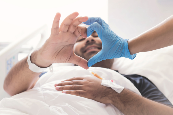 A young man lying in a hospital and his nurse make a love hear with their hands.