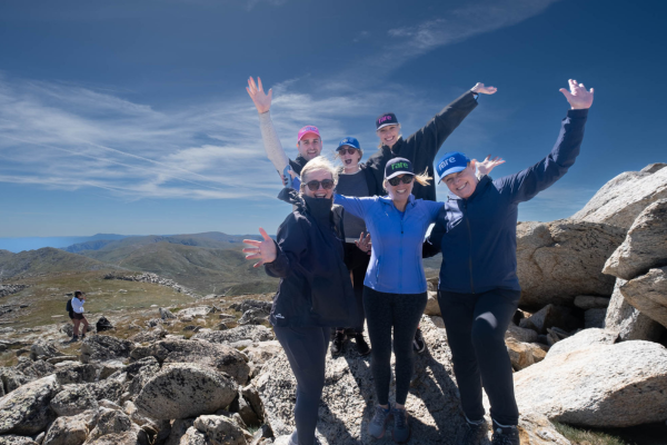 Group of smiling people at the top of Mt Kosciuszko with blue sky and rocky landscape behind them.