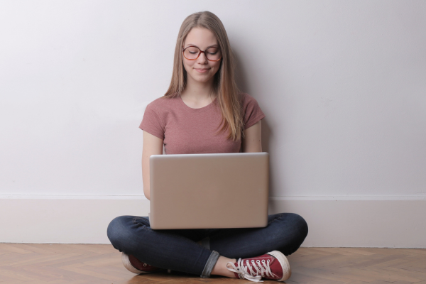 A women wearing a pink shirt and denium jeans sits on the floor with a laptop in her lap.