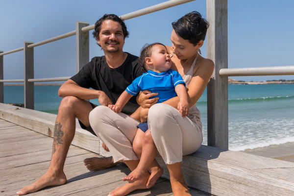 A young man and women sit on a board walk near the beach with their son.