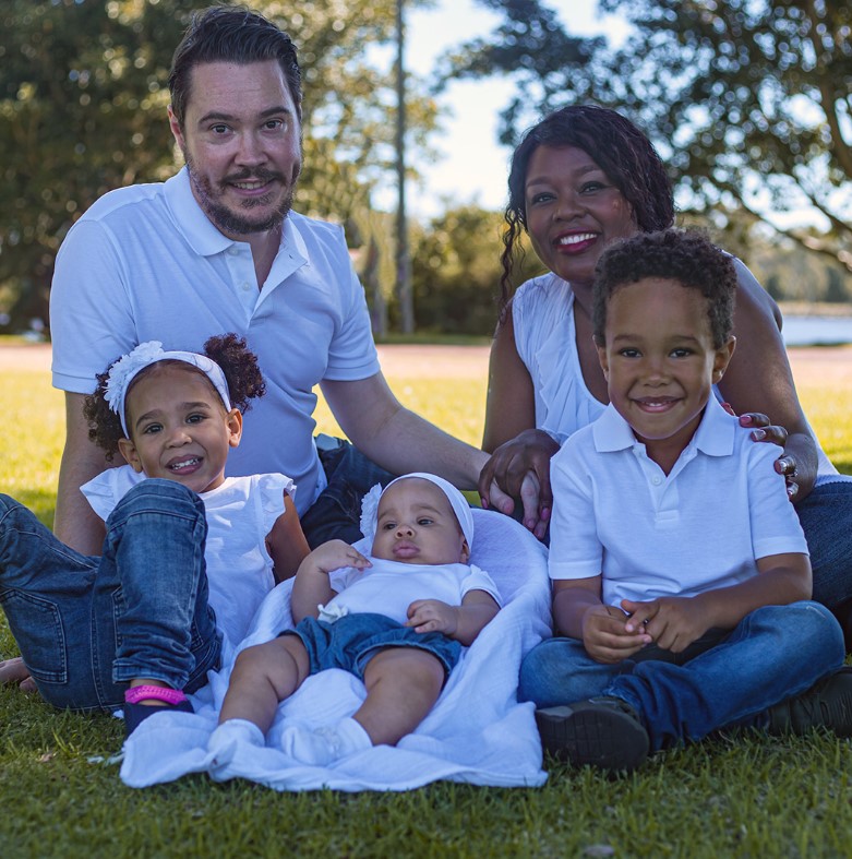 A father and mother smile at the camera with their three children sitting in a park