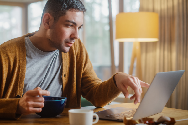 A man sits at his desk whilst he eats breakfast. The man is reading information on his laptop.